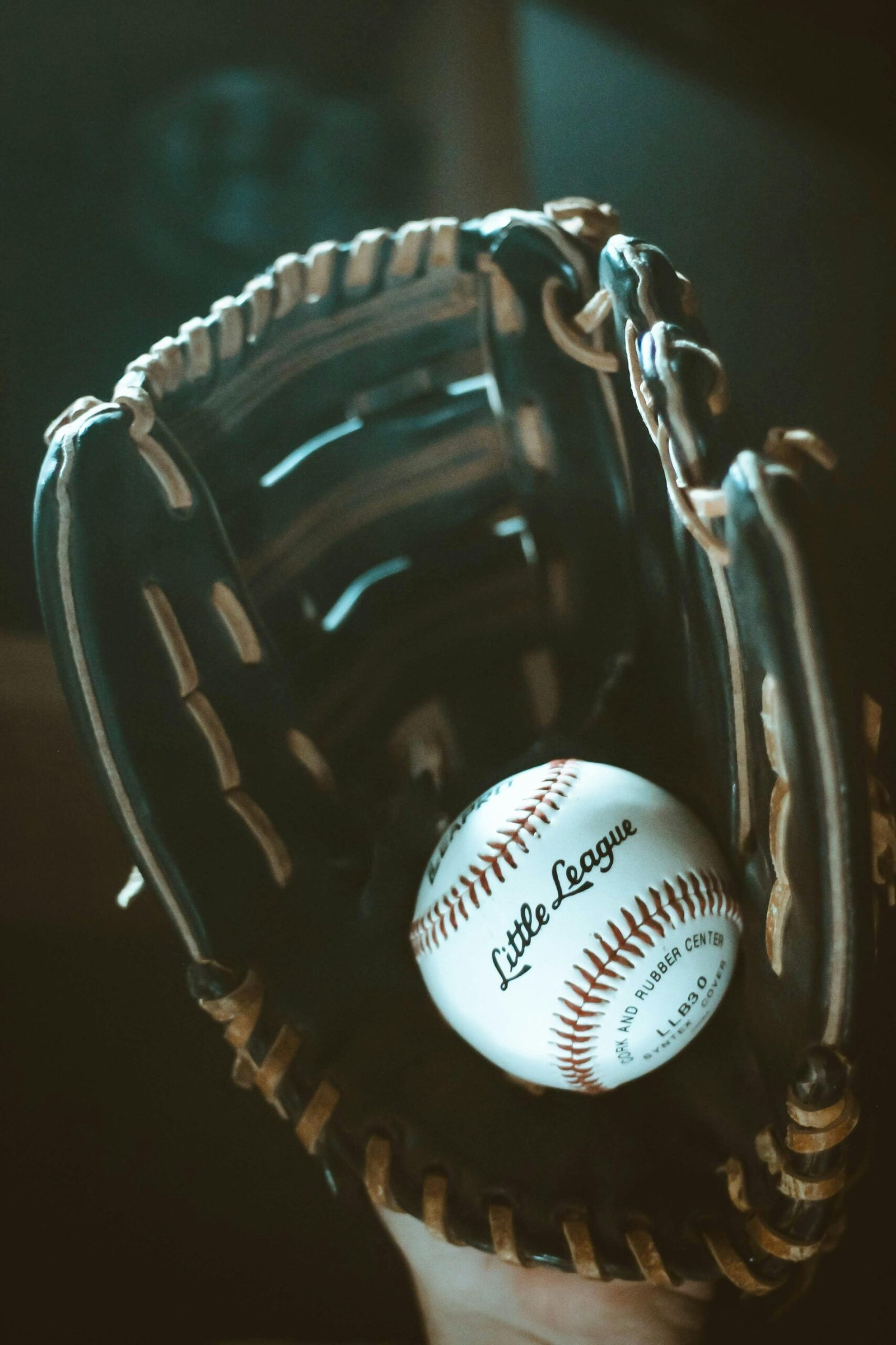 A close-up of a baseball glove holding a "Little League" baseball, with a dark, moody background.